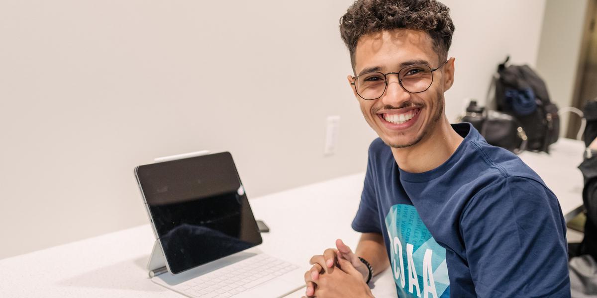 Student smiling at desk with tablet.